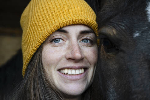 Smiling woman wearing knit hat by piebald horse in stable - KVBF00044