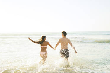 Carefree couple holding hands and running in water at beach - AAZF01663