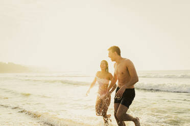 Cheerful young couple playing in water at beach - AAZF01665