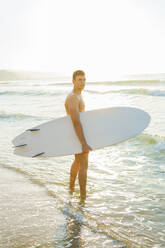 Junger Mann mit Surfbrett im Wasser stehend bei Sonnenuntergang - AAZF01668