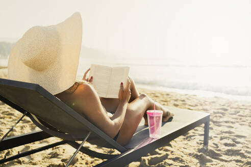 Frau auf Liegestuhl liegend und Buch lesend am Strand - AAZF01674