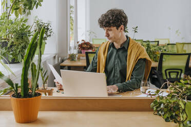 Businessman reading documents near laptop in office - YTF01894