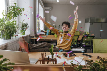 Carefree businessman throwing confetti at desk in office - YTF01921