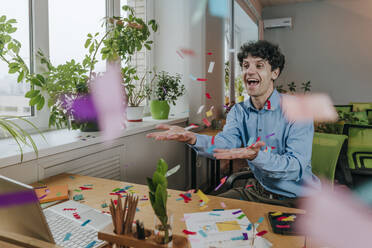 Excited businessman playing with confetti at desk in office - YTF01923