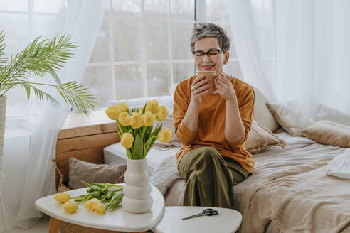 Smiling woman drinking tea near flowers in bedroom at home - YTF01936