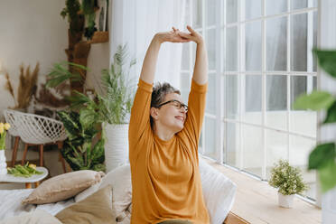 Happy woman stretching arms near window in bedroom at home - YTF01941