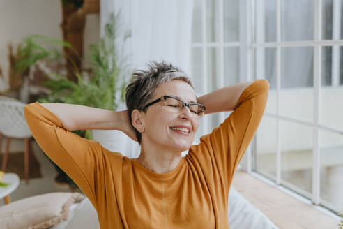 Happy woman with eyes closed and hands behind head at home - YTF01942