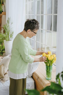 Mature woman arranging tulips in vase on window sill at home - YTF01944