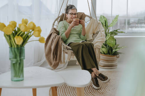 Smiling woman sitting on hanging chair and having tea at home - YTF01948