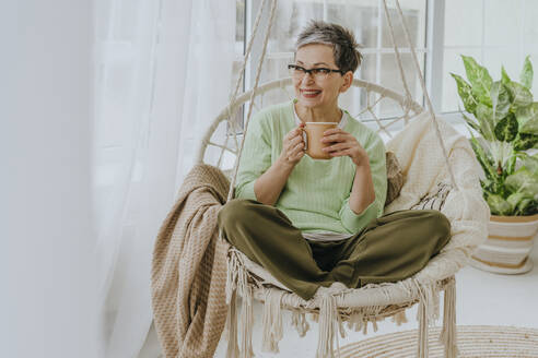 Smiling woman holding mug and sitting on hanging chair at home - YTF01949