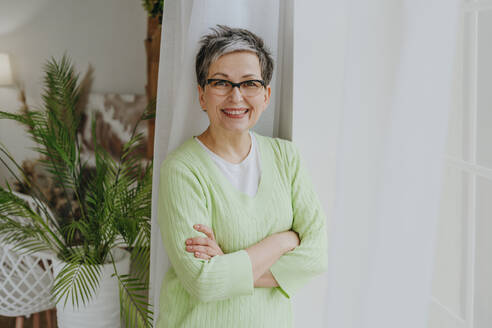 Happy woman with crossed arms standing near window at home - YTF01955