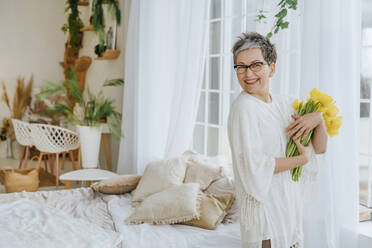 Happy woman holding bunch of tulips near bed at home - YTF01974