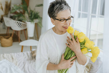 Smiling woman wearing eyeglasses and holding bunch of tulips at home - YTF01975