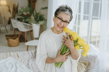 Smiling mature woman holding bunch of tulips at home - YTF01976