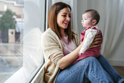 Happy mother with baby girl sitting by window at home - EBBF08803