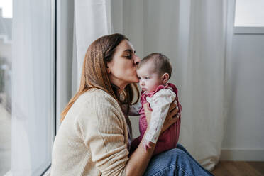 Mutter küsst ihre kleine Tochter, die vor einem Fenster sitzt - EBBF08811
