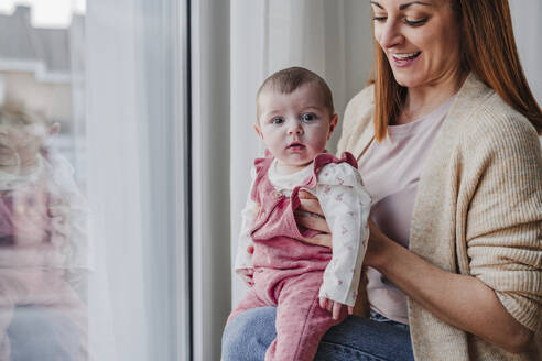 Happy mother with baby daughter sitting by window at home - EBBF08812