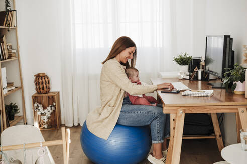 Mutter sitzt mit ihrem kleinen Mädchen auf einem Fitnessball und arbeitet zu Hause im Büro - EBBF08814