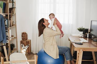 Mother picking up baby girl sitting on fitness ball at home office - EBBF08821