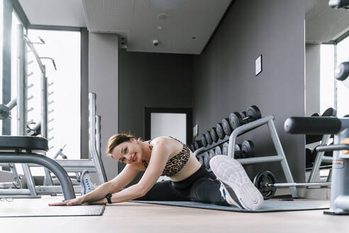 Smiling woman doing stretching exercise on mat in gym - NDEF01590
