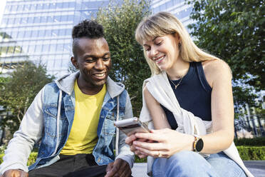 Lächelnde multirassische Freunde, die sich ein Smartphone teilen und in der Nähe eines Gebäudes sitzen - WPEF08477