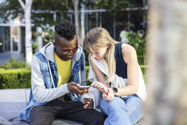 Curious multiracial friends sharing smart phone sitting on bench - WPEF08480