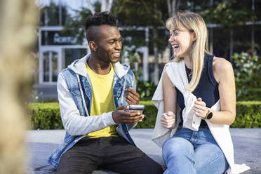 Junge Frau lachend und sitzend mit Freund, der ein Smartphone hält - WPEF08481