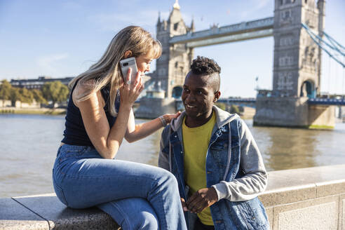 Frau, die mit ihrem Smartphone telefoniert, sitzt an der Wand neben einem Freund mit der Tower Bridge im Hintergrund - WPEF08488