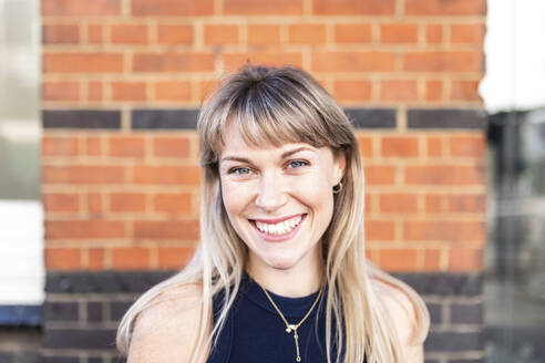 Happy blond woman in front of brick wall - WPEF08509