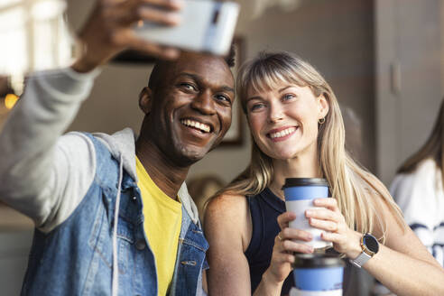 Lächelnder Mann, der ein Selfie mit einem Freund macht, der einen Wegwerfbecher in einem Café hält - WPEF08537