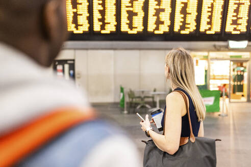 Frau trägt Tasche und liest Zugfahrplan am Bahnhof - WPEF08553