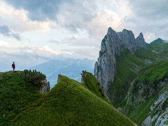 Anonymous hiker stands on a lush green ridge, taking in the breathtaking views of the rugged peaks in the Appenzell region of Switzerland. - ADSF53178
