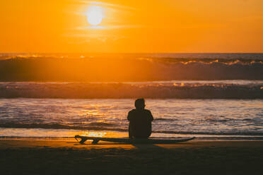 Back view of unrecognizable male surfer in wetsuit sitting on surfboard at seashore and looking at ocean with high water current under orange sky at sunset - ADSF53198