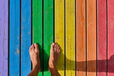 From above of barefoot legs and shadow of anonymous person standing on colorful wooden planks on sunny day - ADSF53201