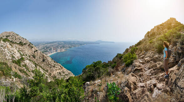 Side view female explorer in casual clothes and trainers holding chain while hiking on rocky slope during summer vacation in Penon de Ifach in Calpe, Alicante, Spain - ADSF53203