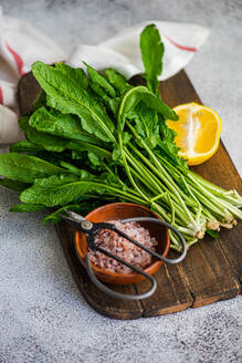 From above of fresh arugula leaves with a slice of lemon and a bowl of salt on a wooden cutting board, accented by kitchen scissors and a striped towel - ADSF53204