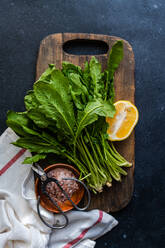 From above of fresh arugula leaves with a slice of lemon and a bowl of salt on a wooden cutting board, accented by kitchen scissors and a striped towel - ADSF53206