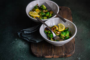 From above of bowl of grilled Brussels sprouts seasoned with garlic and spices, garnished with a slice of lemon, served on a wooden board with a fork - ADSF53207