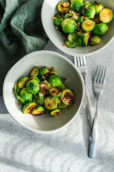 Top view of two bowls filled with barbecued Brussels sprouts flavored with garlic and spices, accompanied by silver forks on a textured tablecloth - ADSF53208