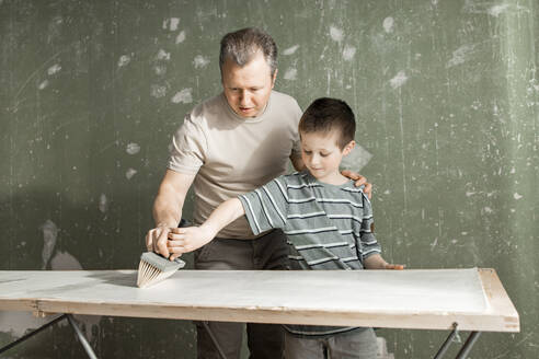 Vater und Sohn tragen gemeinsam Leim auf eine Tapete an der Werkbank auf - ELMF00096