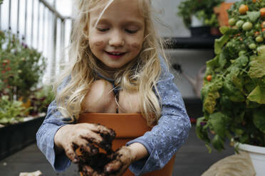 Smiling girl sitting with handful of dirt in pot at balcony - NSTF00014