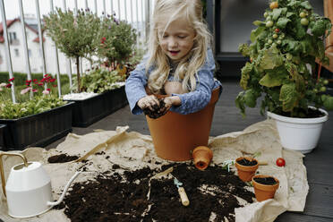 Girl holding dirt and sitting in pot near plants at balcony - NSTF00015
