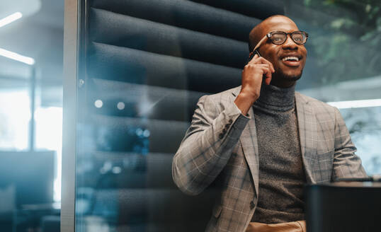 Geschäftsmann in einem Büro, der sein Smartphone benutzt, um zu kommunizieren und über geschäftliche Erfolge zu sprechen. Der Unternehmer trägt eine Brille und wirkt glücklich und professionell in seiner abgelegenen Coworking-Umgebung. - JLPSF31342