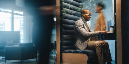 Young man sitting in a busy coworking office, working on a laptop inside a booth. Business people are moving around in the shared workspace. Professional, happy, and successful workplace. - JLPSF31343