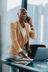African businesswoman in a corporate office uses her smartphone to talk and discuss work. She stands by her desk with a laptop, displaying professionalism, success and happiness. - JLPSF31359