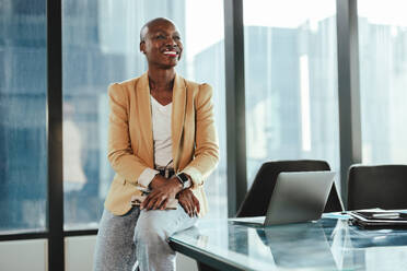 Selbstbewusste afrikanische Geschäftsfrau in einem Büro, an einem Tisch sitzend. Sie ist glücklich und erfolgreich, lächelt, während sie den Blick abwendet. Ihre Professionalität und ihr Unternehmergeist tragen zum Erfolg ihres Unternehmens bei. - JLPSF31362