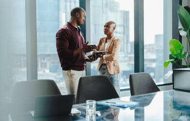 Two professionals standing in a boardroom together, discussing business and planning corporate deals. Teamwork, technology, and success are evident as they utilize a tablet to collaborate. - JLPSF31374