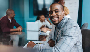 Ein professioneller Geschäftsmann in einem eleganten Anzug strahlt mit einem warmen Lächeln während einer Teambesprechung in einer modernen Büroumgebung Selbstvertrauen aus und verkörpert damit Führungsqualitäten und Positivität. - JLPSF31390
