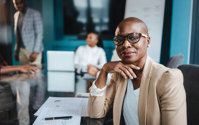 Eine afrikanische Geschäftsfrau mit Brille leitet selbstbewusst eine Sitzung in einem Sitzungssaal mit ihren Kollegen, die Dokumente und Berichte besprechen und auf den Erfolg im Büro hinarbeiten. - JLPSF31391