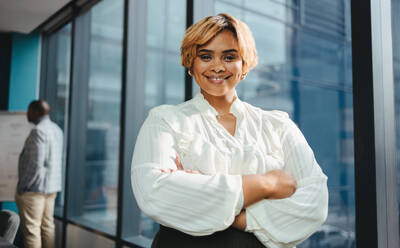 Happy and successful black businesswoman stands in a boardroom office. She has her arms crossed, smiling and looking directly at the camera. With a professional demeanor, she exudes confidence in her workplace. - JLPSF31396